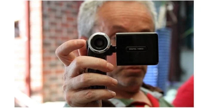 Man holding a video camera pointed directly at the viewer with face obscured.